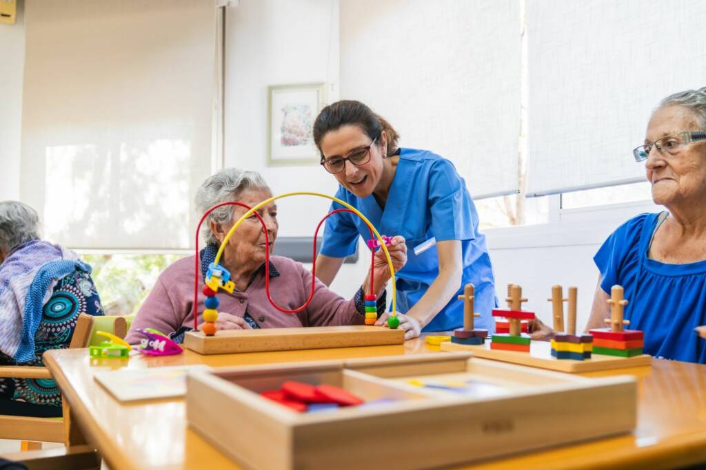 Pflegefachkraft assistiert Seniorinnen bei ergotherapeutischen Übungen mit bunten Motorikschleifen und Holzspielzeug in einer Pflegeeinrichtung.
