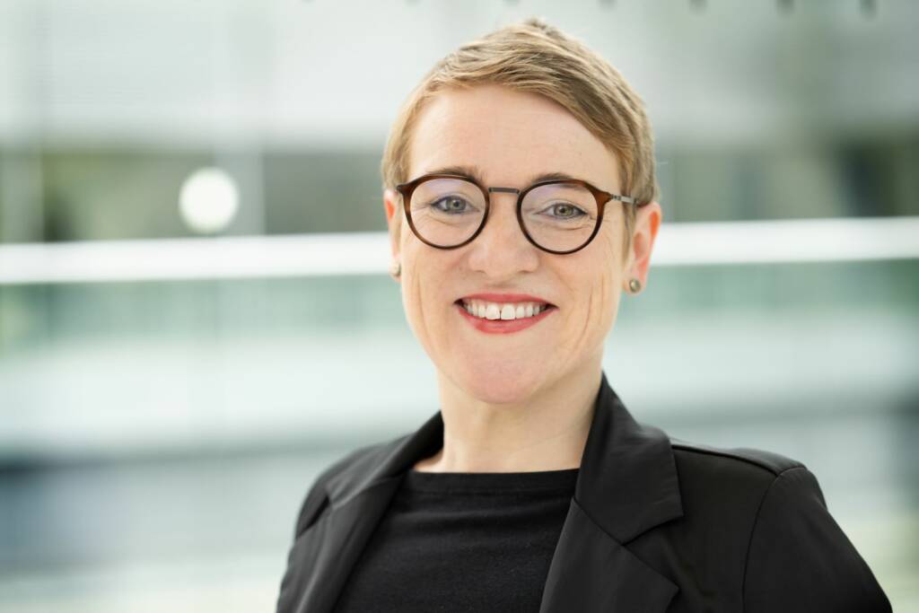 Professionelles Porträt einer Person mit Brille in schwarzem Blazer vor unscharfem Bürogebäudehintergrund mit natürlichem Licht.