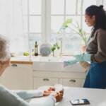 Rare view of elderly lady with gray hair looking at african american housekeeping volunteer helping to clean house, washing kitchen sink with detergent, wearing raisin gloves, talking to each other